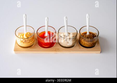 Four glass sauce bowls with white ceramic spoons on natural wooden bamboo base, colorful condiment serving set for kitchen styling and culinary produc Stock Photo