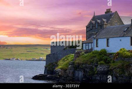 Historic stone buildings perched on dramatic rocky cliffs overlooking the sea at vivid pink and orange sunset in Lerwick, Shetland Islands, Scotland Stock Photo