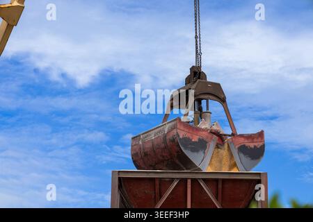 A heavy industrial crane clamshell grab bucket suspended by metal chains above a rusty cargo container at a port terminal against a bright blue sky wi Stock Photo