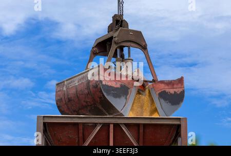 A heavy industrial crane clamshell grab bucket suspended by metal chains above a rusty cargo container at a port terminal against a bright blue sky wi Stock Photo
