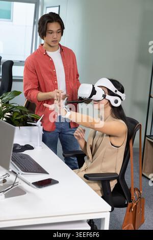 Woman wearing VR headset reaching, engaging UI floating in office for training as man guiding Stock Photo