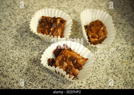 Three homemade chocolate chip brownies, each nestled in a white paper cup, sit on a speckled countertop, showcasing their rich, gooey texture. Stock Photo