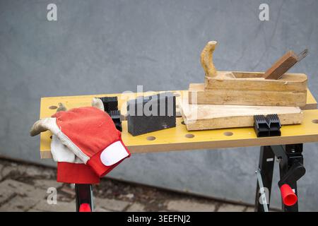 A wooden hand plane, sandpaper block, work gloves, and clamps are arranged on a portable workbench, ready for carpentry tasks Stock Photo