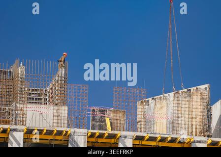 Construction worker in a hard hat overseeing a large precast concrete wall panel being lifted by a crane on a high-rise building site, showcasing the active phase of urban infrastructure development Stock Photo