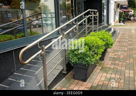 Green decorative bushes in pots decorate a modern entrance to the building with a ramp for the disabled and stainless steel handrails. Stock Photo