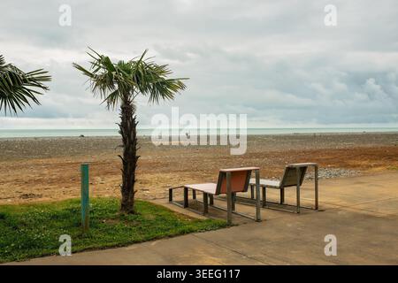 Two sunbed-style benches installed on a seaside boulevard in front of a pebble beach with palm trees against the Black Sea under dramatic clouds. Stock Photo