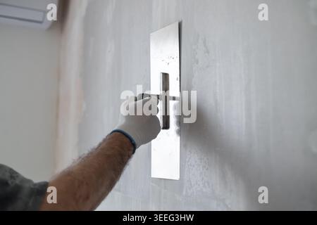 Finishing contractor using stainless steel trowel to apply a smooth layer of plaster on interior wall. Clean workspace with bright lighting and minimal distractions visible in background. Stock Photo