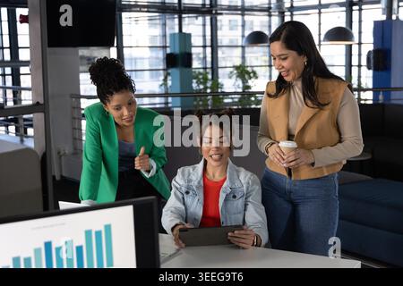 Diverse female team collaborating in workwear around table in office lounge with tablet and coffee Stock Photo