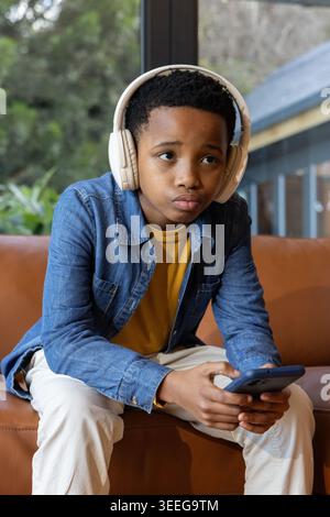 Preteen African American boy sitting on couch near window in denim jacket with headset using phone Stock Photo