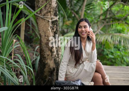 A horizontal medium shot captures a smiling Thai woman talking on her smartphone while sitting in a lush jungle setting in Phuket, Thailand. Dressed i Stock Photo