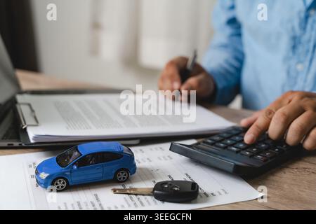 Hand using calculator for car finance loan payment calculation with blue toy car model, key and financial documents on desk, representing auto financi Stock Photo