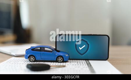 Blue toy car on insurance document beside smartphone with protection icon, representing car insurance coverage, vehicle protection, digital claim serv Stock Photo