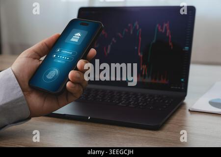 Hand holding smartphone with mobile online banking login screen in front of laptop displaying financial trading chart, suitable for fintech security, Stock Photo