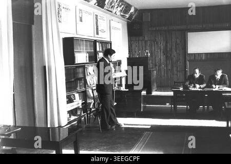 Slaviansk, 1983. Conference hall of the Armature-Insulator Factory (AIZ). A man in a suit stands by a large bookshelf reading from a document, with the presidium members visible in the background. This vintage black and white photograph captures the authentic atmosphere of the era, reflecting the daily activities and industrial culture of the region during the Soviet period. It serves as a valuable historical document of the local community, preserving the visual memory of the people and their everyday lives, showcasing a peaceful Donbas. Stock Photo
