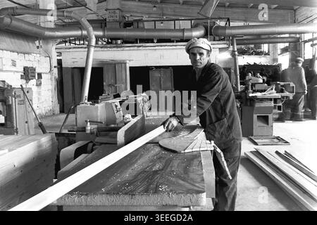 Slaviansk, 1983. Inside a workshop at the Armature-Insulator Plant (AIZ). A male worker stands at a large woodworking machine, carefully guiding a long wooden board through the planer while looking directly at the camera. This vintage black and white photograph captures the authentic atmosphere of the era, reflecting the daily activities and industrial culture of the region during the Soviet period. It serves as a valuable historical document of the local community, preserving the visual memory of the people, showcasing a peaceful Donbas. Stock Photo
