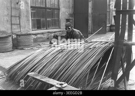 Slaviansk, 1983. Factory territory of the Armature-Insulator Plant (AIZ). A male worker wearing protective goggles is actively managing a massive, heavy coil of thick steel wire outdoors near a brick industrial building. This vintage black and white photograph captures the authentic atmosphere of the era, reflecting the daily activities and industrial culture of the region during the Soviet period. It serves as a valuable historical document of the local community, preserving the visual memory of the people, showcasing a peaceful Donbas. Stock Photo
