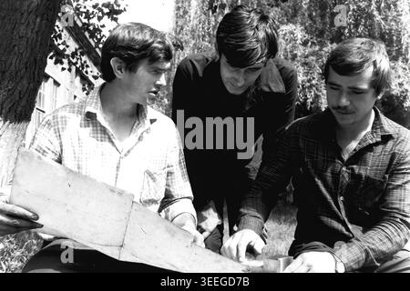 Slavyansk, 1983. Territory of the Armature-Insulator Plant (AIZ). Black and white archival photograph capturing a fascinating historic scene. Three focused male engineers sit outdoors under the refreshing shade of a leafy tree. They are deeply engaged in reviewing a large, unfolded technical blueprint or paper schematic, collaborating closely on the plant's essential manufacturing or construction plans. This detailed image preserves a genuine fragment of Soviet industrial history, showcasing life before the war and peaceful Donbas. Stock Photo