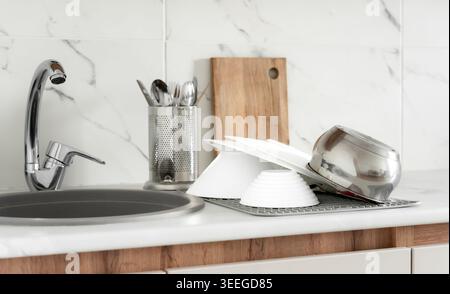 Modern kitchen sink area with washed dishes, cookware and drying rack on white countertop with marble backsplash. Stock Photo