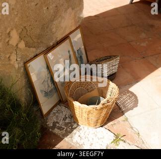 Antique French flea market with vintage wicker baskets and framed art by sunlit stone wall. Rustic outdoor display of authentic Mediterranean Stock Photo