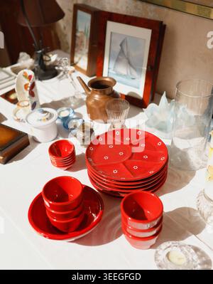 Antique French flea market treasures with red ceramic plates and vintage kitchenware on white table. Vibrant Provence brocante display of copper jugs Stock Photo