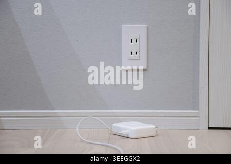 A white electrical outlet on a light grey interior wall, with a white power charger and cable on the wooden floor below. Modern domestic scene evoking home tech and daily living. Stock Photo