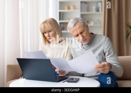 Concerned senior couple reading financial papers at home, looking worried Stock Photo