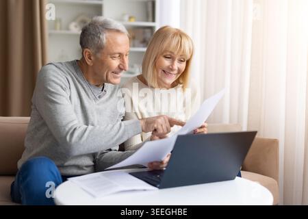 Mature couple checking financial documents with laptop at home, discussing bills Stock Photo