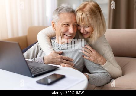 Loving mature woman hugging smiling husband while sitting near laptop at home Stock Photo