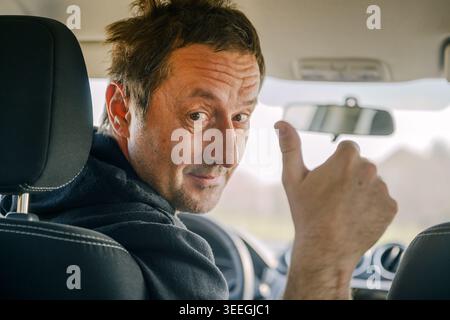 Smiling driver giving thumbs up inside car, positive gesture and confident mood during travel, selective focus Stock Photo