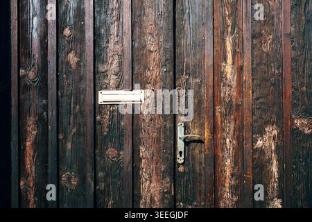 Old vertical wooden door in deep brown tones with weathered grain, brass mail slot and vintage handle. Rich texture highlighted by soft side light. Stock Photo