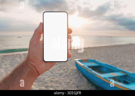 Hand holding smartphone with blank screen at beach with small blue boat and sunset in background, outdoor app mockup or web design. Selective focus. Stock Photo