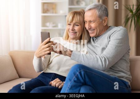 Smiling mature couple using smartphone together at home, enjoying digital communication Stock Photo