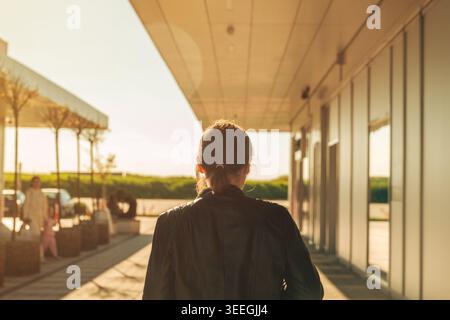 A woman seen from behind walks along a modern terminal walkway at sunset, evoking travel, quiet reflection and transit. Warm light and contemporary ar Stock Photo