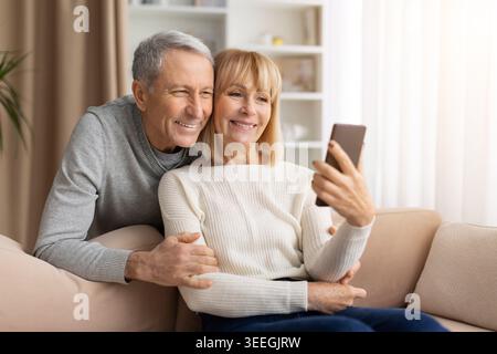 Affectionate mature couple relaxing on sofa at home with smartphone Stock Photo