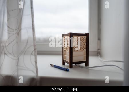 A small Kumiko wooden decorative lamp sits on a bright windowsill beside a blue pen and sheer curtain, creating a calm, cozy interior scene. Stock Photo