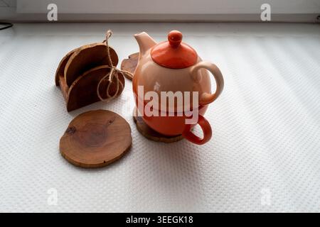 Small stacked orange teapot and matching cup sit on round wooden coasters beside a wooden coaster holder on a white textured surface. Stock Photo
