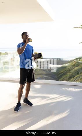African American man in blue athletic shirt drinking from sports bottle on terrace holding mat, lid. Balcony, oceanfront, fitness, outdoors, sunny, hy Stock Photo