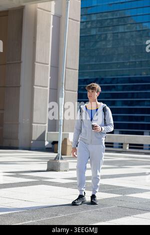 Man walking across tiled urban plaza in grey tracksuit carrying coffee cup and black backpack. Pedestrian, cityscape, outdoor, contemporary, minimalis Stock Photo