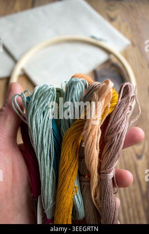 Hand holding colorful embroidery floss threads with wooden embroidery hoop and fabric on rustic wooden background. Handmade craft and needlework conce Stock Photo