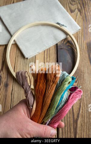 Hand holding colorful embroidery floss threads with wooden embroidery hoop and fabric on rustic wooden background. Handmade craft and needlework conce Stock Photo