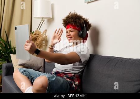 A cheerful teen sits on the couch, using headphones and a tablet to connect with friends. Stock Photo