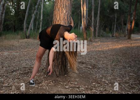 Young woman bending backwards, stretching her body and leaning on a tree trunk in a natural outdoor setting at sunset Stock Photo