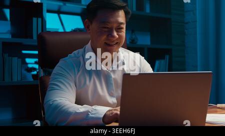 Happy motivated middle-aged Korean business man working laptop in night office Chinese businessman typing computer managing online job task startup Stock Photo
