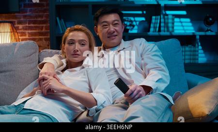 Multiracial relaxed couple watching tv searching television program talking resting relaxing lying sitting on couch sofa multiethnic family watch Stock Photo