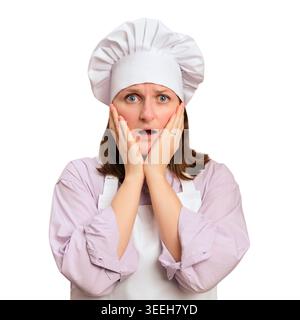 Surprised woman chef wearing hat and apron looking at disaster in kitchen. Shocked cook with hands on cheek. Emotional reaction to recipe mistake. Cul Stock Photo