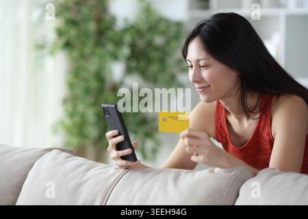 Satisfied asian woman paying online using credit card and cell phone at home Stock Photo