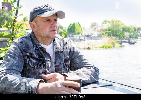 A middle-aged man relaxing at a waterfront terrace, wearing a grey denim jacket and baseball cap, holding a coffee cup and an electronic cigarette Stock Photo