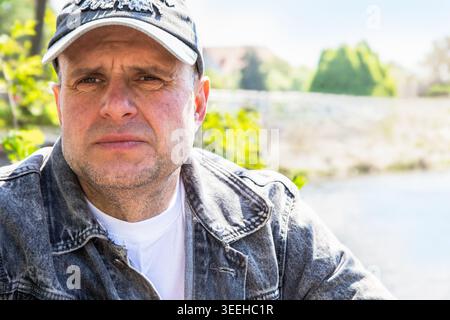 Thoughtful middle-aged man with a serious expression wearing casual denim clothes, sitting outside on a bright sunny day with soft bokeh Stock Photo