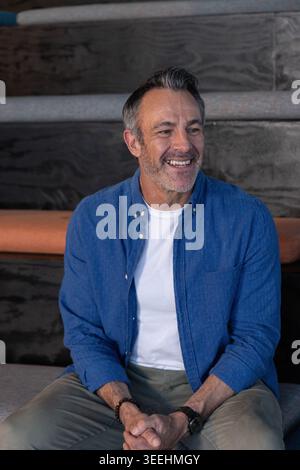 Middle-aged man sitting on tiered bench smiling with hands clasped in shirt with orange pad behind Stock Photo