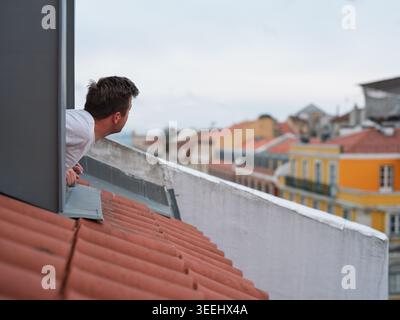 Title: Curious man leaning out of a roof window looking at the urban cityscape Stock Photo